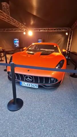 A bright orange sports car with black accents is displayed indoors under a tent-like structure with spotlights. The car is a Mercedes-Benz and has a German license plate that reads "GT XX 5479". The vehicle is cordoned off with stanchions and velvet ropes. There are digital displays in the background. The camera is moving slightly, giving a panning shot of the car.