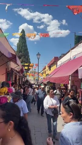 The video shows a view looking down a crowded outdoor market street. Buildings on both sides of the street have awnings that extend over the sidewalk. Vendors are selling wares under the awnings. Colorful banners are strung across the street overhead. People are walking in both directions. The sky is blue with some white clouds. There is a tall thin tree visible in the distance. The music playing is loud and upbeat.