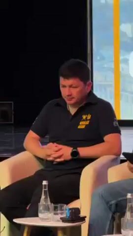 The video shows a medium shot of a man seated in a light-colored armchair. He is wearing a black polo shirt with yellow text that says “MYKOLAIV REGION” and a design. He also wears a black smartwatch on his left wrist. His arms are folded in his lap. He is looking to his left with a neutral expression. To his right, part of another person seated in a similar chair is visible. There is a clear plastic water bottle on a surface to the left of the man. The background is a dark stage with a blurred yellow and white screen.