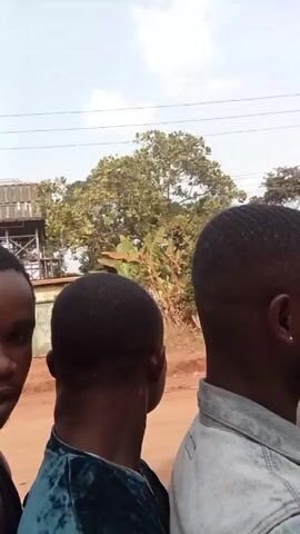 Three people are seated on what appears to be a motorcycle or similar small vehicle, moving along a dirt road. The person in the middle wears a denim jacket and has a beard. The person in front is wearing a blue-green velvety shirt. The person in the back has a tan shirt. The camera is positioned behind and to the side of the group, capturing their backs and profiles. The background shows dry grassy vegetation, trees, and palm trees under a bright sky. The sound of wind is prominent.