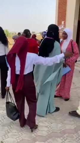 A group of people, mostly young, are gathered outside a brick building. Many are wearing traditional clothing or head coverings. Some are greeting each other with handshakes. They are walking around and talking. There is a camera tripod visible. The general atmosphere seems lively and possibly celebratory.