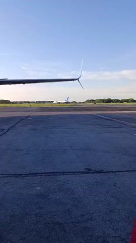 The video captures an airport tarmac scene from the perspective of an aircraft. The wing and winglet of the camera's plane are visible on the left side of the frame. The tarmac is dark grey with a prominent red painted line visible on the right. In the middle distance, several other airplanes are seen: an initial white aircraft with blue markings, followed by a dark-colored aircraft with red accents, and a mostly red aircraft further behind it. These planes appear to be parked or slowly taxiing. A ground crew member wearing a high-visibility yellow and blue jacket briefly walks into the frame from the right. The sky above is clear and bright blue with some scattered white clouds, indicating daytime conditions, possibly morning or late afternoon. The setting is an active airfield at Bolshoye Savino, Russia. No clear text is discernible on the aircraft or in the environment.