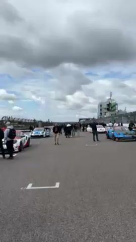 The video shows a series of racing cars lined up on a track, with people milling around and observing. There is a blue and white car with the number 3 on it, with mechanics making adjustments to the car. The track is gray asphalt with white painted lines. There is a grandstand and control tower in the background, and flags are visible in the distance. The sky is overcast.