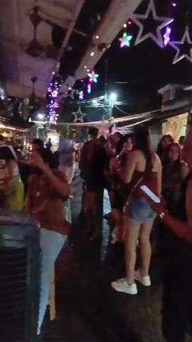The video shows an outdoor night scene with a crowd of people gathered together under what appears to be an open roof. Decorations including star-shaped lights and colorful string lights are strung above. People are wearing various casual outfits, and some are holding drinks and phones. Some individuals are taking pictures or videos with their phones. The ground appears wet, possibly from a recent cleaning or rain. There is a boxy object, possibly an air conditioning unit, visible in the foreground on the left. The atmosphere appears lively and festive, suggesting a social gathering or event.