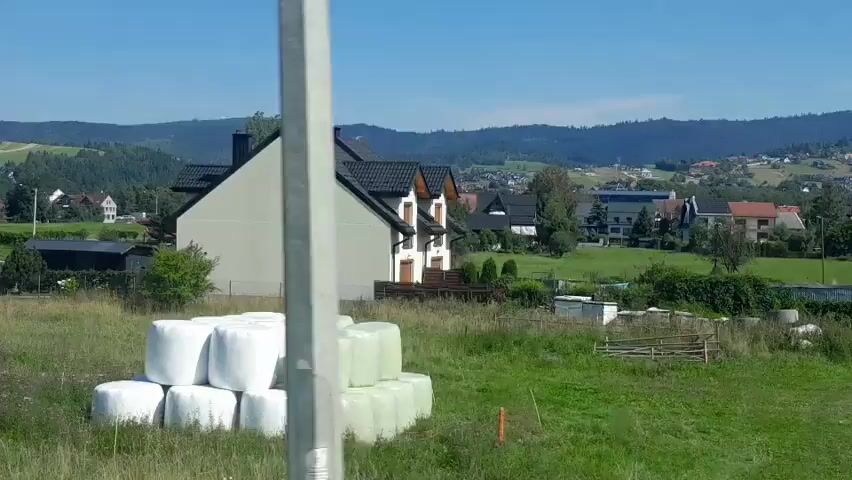 A drone camera pans right over a grassy field in a rural area, showcasing a stack of white hay bales in the foreground. Beyond the field, several houses are visible, some with dark roofs, nestled amongst trees and rolling hills. The sky is clear and blue, indicating a sunny day. The drone appears to be at a moderate altitude, offering a panoramic view of the landscape.