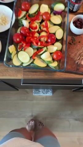 The video shows a top-down view of a person's legs and feet standing over a wooden floor. In the background, a counter holds several bowls and a glass baking dish. The baking dish contains sliced zucchini, cherry tomatoes, red bell pepper, and what appear to be red onions. One white bowl holds chopped garlic, and another white bowl contains dark olives. A wooden cutting board on the right side of the counter has some green capers and what looks like raw meat. The person's legs are visible, wearing light gray shorts. Their feet are bare, and one foot has painted toenails. The scene appears to be a kitchen, with the person preparing ingredients for a meal.