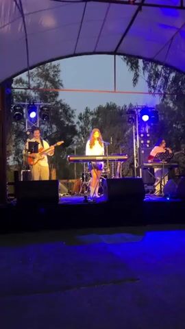 The video shows a live musical performance taking place on an outdoor stage. The stage is covered by a white tent-like structure. Three musicians are visible on stage. On the left, a male musician wearing a hat and a light-colored shirt is playing a guitar. In the center, a female musician wearing a white top and a short skirt is singing and playing a keyboard. On the right, another musician, likely female, is playing a keyboard and possibly singing. The stage is lit with blue and white lights, and there are speakers in the foreground. Trees and the evening sky are visible in the background.