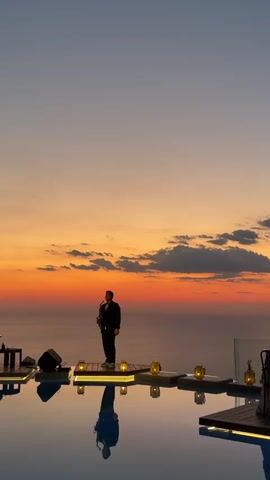 A man in a suit plays the saxophone on a raised platform by a swimming pool during sunset. He is reflected in the pool's water. In the background, a band setup is visible to the left, and two people are seated at a table to the left, facing the ocean. The sky is a gradient of orange and purple with clouds. The setting appears to be a luxury resort or restaurant overlooking the ocean.