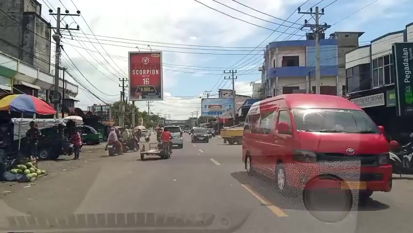 A street scene in a town or city. The day is bright and cloudy. In the foreground, the dashboard of a vehicle is visible, indicating the video is from inside a car. The road is paved and has yellow lane markings. There is moderate traffic, consisting of cars, motorcycles, and a few three-wheeled vehicles. People are seen riding motorcycles and walking on the sidewalks. Buildings line the street, some appear to be shops or businesses, with awnings and advertisements visible. A large billboard for "Red Scorpion" cigarettes is prominently displayed. Power lines crisscross the sky. On the left side of the road, there are market stalls with produce, possibly watermelons. The overall impression is of a busy, typical day in a commercial area.