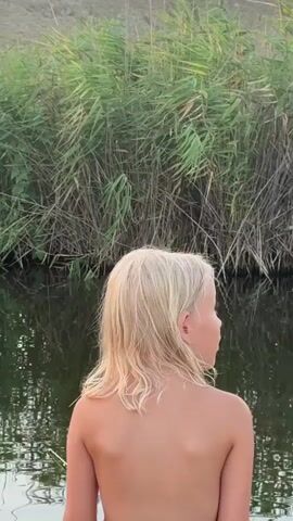 This media file captures a serene moment by a body of water, likely a canal or a slow-moving river, in the Урожайновское сельское поселение, Ukraine. The scene is set during the golden hour, with the sky transitioning through soft hues of orange and yellow, indicating either sunrise or sunset. Tall, reedy grasses line the opposite bank of the water, creating a natural, verdant backdrop.

The primary subject is a young child with blonde, wavy hair, seen from the back. Initially, the child appears to be sitting directly by the water's edge, their bare back exposed. In subsequent frames, the child is seated on a small, white, child-sized chair with colorful stickers on its backrest. The child is wearing dark shorts. To the right of the child, a fishing rod is visible, propped up near the water.

To the left of the child, a large, rolled-up pink mat and a small pile of items, including a water bottle and some green objects, are present on the sandy bank. The overall atmosphere is peaceful and suggests a leisurely outdoor activity, possibly fishing or simply enjoying the natural surroundings as the day draws to a close or begins. The gentle reflection of the reeds and sky on the water's surface adds to the tranquil mood. There is no visible text within the media.