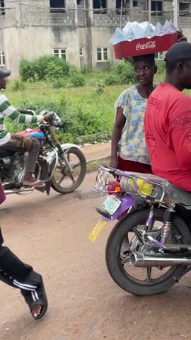 The video depicts a street scene in what appears to be a developing country. A woman carries a red Coca-Cola crate filled with bottled drinks on her head.  A man sits on a parked motorcycle eating something from a small plastic bag. Another motorcycle, with a man in a green and white striped shirt riding it, passes. A table is laden with sunglasses displayed for sale. There is a glass display case with food items on top of the table. A man sits on a blue stool behind the table, possibly the vendor of the goods. The setting appears to be a somewhat rural or suburban area with buildings and greenery visible in the background. There are puddles on the dirt road.