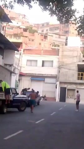 A group of people are gathered on a street in what appears to be a hillside urban environment. On the left, a person in a neon green shirt and dark shorts is on the back of a pickup truck. In the center, a dark-colored pickup truck is stopped in the street, and several people are around it. One person, shirtless and wearing dark shorts, stands in front of the truck. Another person, also shirtless and wearing dark shorts, appears to be leaning into the driver's side window. Two other individuals are near the truck. Further down the street, a person in a pink shirt and dark shorts is walking away from the camera. Buildings are visible on either side of the street, with more buildings cascading up the hillside in the background. Trees with green leaves are visible in the foreground, partially obscuring the sky. The lighting suggests it is daytime, possibly late afternoon.