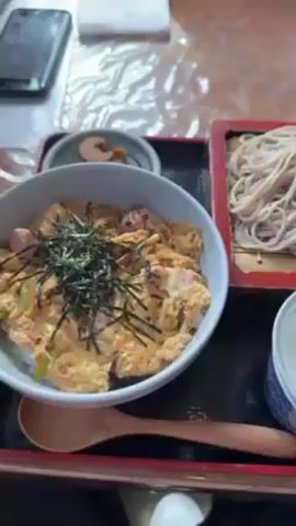 The video shows a black tray with a bowl of Japanese rice dish topped with seaweed, a wooden spoon, a serving of soba noodles in a red tray, a bowl of grated radish, wasabi, and sliced green onions, and a small dish of pickled ginger. Another similar set of soba noodles with the radish, wasabi, green onions, and pickled ginger is also visible. The video shows a hand briefly moving the tray. The background shows a blurred table and a phone.