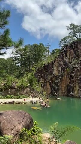 The video shows a serene outdoor scene with a crystal-clear emerald green lake or pond. Tall, rugged rock formations with a layered texture rise dramatically on the right side of the frame, partially covered in vegetation. The landscape is lush and green, with dense trees and foliage surrounding the water. Several people are visible near the water's edge and on a makeshift raft in the lake. One person is standing on a rocky outcrop near the shore, while a group of individuals are gathered on the raft, some appearing to be relaxing or preparing to swim. The sky above is a vibrant blue, dotted with fluffy white clouds, suggesting a sunny day. The overall atmosphere is peaceful and suggests a recreational or natural escape.