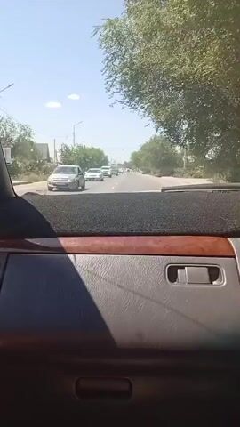 The video shows a view from inside a car traveling on a road. The road is paved and has lane markings. There are other cars on the road, including a white SUV in the foreground and several other vehicles further down the road. Trees and vegetation line the sides of the road, and there are some buildings visible in the distance. The sky is clear and blue, indicating a sunny day. The interior of the car is partially visible, including the dashboard with a dark mat and a wood-grain trim. The perspective suggests the car is moving forward.
