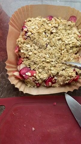 A high-angle shot shows a person’s hands using a spoon to press oats into a fruit crumble. The crumble consists of oats and red-skinned fruit, possibly apples or rhubarb, and is contained within a square baking pan lined with parchment paper. A red cutting board and a silver knife are visible in the bottom of the frame. The person is wearing a purple long sleeve shirt. The background is a gray countertop.