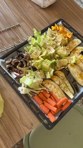 A close-up video shows a baking tray filled with a variety of chopped and seasoned vegetables, ready for cooking. The tray, lined with parchment paper, rests on a light-colored wooden surface, likely a kitchen counter.

The vegetables are arranged in distinct sections:
- On the left, there are mushrooms and what appears to be chopped leeks or spring onions.
- In the center, a large pile of roughly chopped green cabbage is visible, seasoned with spices.
- To the right of the cabbage, several slices of eggplant are arranged, also seasoned.
- In the bottom right corner, a section of chopped carrots is present.
- In the top right corner, diced yellow bell peppers complete the colorful assortment.

Most of the vegetables are sprinkled with visible spices, including what looks like black pepper and a yellowish seasoning, possibly turmeric or curry powder. An empty oven rack is visible in the background, suggesting the vegetables are prepared for roasting or baking. No people or text are visible in the video.