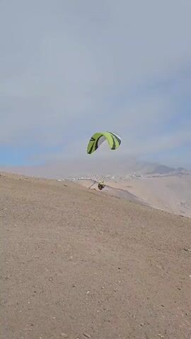 A person in yellow and black gear is paragliding from a hill. The paraglider is green and white with the word "TEQUILA" visible on its surface. The background shows a valley with mountains and a town near the ocean. The sky is partially cloudy. The camera is positioned on the hill and follows the paraglider as it ascends.