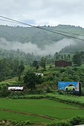 This is a landscape video of a mountainous region covered in fog. In the foreground, there are green agricultural fields. Further up the slopes, there are trees and small houses. There is a billboard visible on the right side of the screen. The video pans across the scenery.