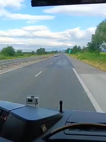 The video shows a view from inside a vehicle, looking forward through the windshield. The road ahead is a highway with multiple lanes. There are other vehicles on the highway in the distance. The sky is cloudy, and there are trees and fields on either side of the road. On the dashboard of the vehicle, there is a small electronic device with a screen and buttons, and a black object that appears to be a stand or holder. The steering wheel is partially visible.