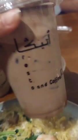 A close-up shot focuses on a clear plastic cup filled with a brown liquid and ice, topped with a layer of brown powder. The cup has black text printed on it, including Arabic script and the words "resto and Coffee." A hand is holding the cup from the top, with fingers visible. In the background, a bowl of noodles with vegetables and shrimp is visible, suggesting a meal setting. The lighting is warm and diffused. The video appears to be a short, casual recording of a beverage and food.