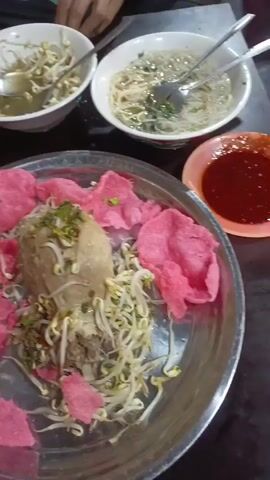 The video shows a close-up of a plate of food, likely bakso, a type of Indonesian meatball soup. The plate is large and silver, and it contains a very large meatball on top of a bed of bean sprouts and what appears to be some minced meat. There are also pink crackers, possibly kerupuk, scattered around the edges of the plate. In the background, out of focus, are two bowls of soup, also containing bean sprouts and noodles, and what looks like a small dish of chili sauce. A hand with a spoon is visible reaching towards one of the bowls of soup. The overall setting appears to be a casual dining environment, possibly a restaurant or food stall, with a dark background.