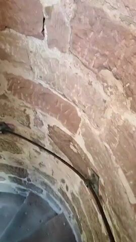 The video shows a spiral staircase made of stone with a cable railing. The walls are constructed of rough-hewn stones. The camera is moving downwards, giving a view of the steps and the cable. There is a window at the top of the staircase, with light coming through it.