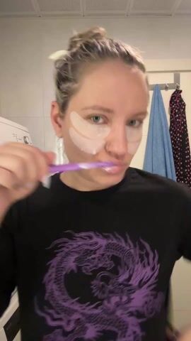 A woman stands in a bathroom brushing her teeth. She wears a black t-shirt with a purple dragon design and has two white under-eye patches on her face. Her blonde hair is pulled up in a messy bun. The bathroom is white-tiled with a washing machine visible on the left. A towel and robe hang on a rack behind her. The camera angle is slightly high, focused on the woman’s face as she brushes her teeth with a purple toothbrush.