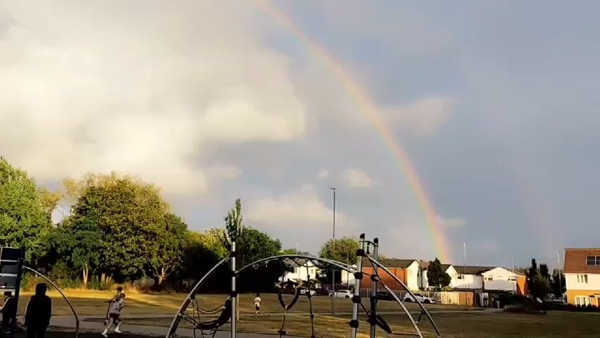 The video pans across a park scene with a rainbow visible in the sky. In the foreground, there are playground structures and several people, some of whom appear to be children, are engaged in activities. In the background, there are trees and residential buildings. The sky is cloudy with patches of sunlight, and the rainbow arcs across the frame.