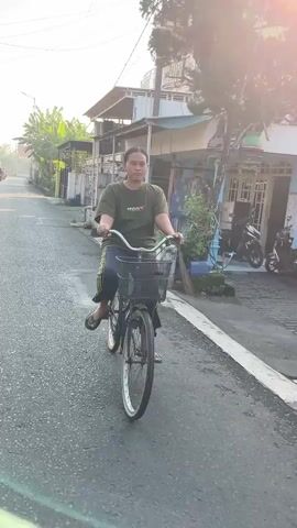A young person is riding a bicycle down a paved street. The person is wearing a green t-shirt and dark pants with yellow stripes. They are facing forward and appear to be looking towards the camera. The bicycle is black with a basket on the front. The street is lined with houses and some vegetation. There is a motorcycle parked on the right side of the street. The sun is shining, casting a warm glow on the scene.