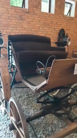 The video shows an antique horse-drawn carriage displayed indoors, likely in a museum or exhibition. The carriage is made of wood and metal, with dark upholstered seats. It has large spoked wheels. The background consists of a brick wall with several open windows. The lighting is moderate. There are no people visible in the frame. The camera is stationary and positioned at a medium distance, focusing on the carriage.
