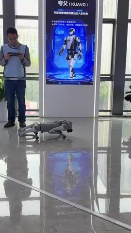 A man stands in an open room, holding a phone in his hands and looking at the screen. He's wearing jeans, a grey shirt, and a light-blue safety vest. Adjacent to him is a large screen showing a humanoid robot. Closer to the camera, a quadruped robot, resembling a dog, is crawling on the polished floor, reflecting the images of the robots from the nearby screen. A potted plant stands near the large window to the right.