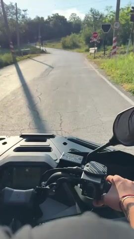 The video captures a first-person perspective of someone riding an ATV on a paved road in Boltata, Bulgaria. The scene is set in a rural area with lush green trees and vegetation lining the road. The sun is shining, indicating it's daytime, and the weather appears to be pleasant.

The ATV's handlebars and dashboard are prominently visible in the foreground, with a person's hands operating the controls. The rider is wearing a black t-shirt, and a bracelet with red and white beads is visible on their wrist. The ATV itself is black and appears to be a modern model.

The road leads to a railway crossing. A red octagonal "STOP" sign is displayed above a white X-shaped sign with red stripes, indicating the presence of the railway tracks. A metal guardrail runs along the side of the road near the crossing. The railway tracks are visible crossing the road, and in the distance, the tracks curve to the left, disappearing into the trees.

The overall mood is one of exploration and enjoyment of the outdoors, with the rider navigating through a scenic, rural Bulgarian landscape. The video appears to be a personal recording of a recreational ATV ride. No other people are clearly visible besides the rider's hands and a brief glimpse of the rider's head from behind. No other text is clearly legible besides the "STOP" sign.