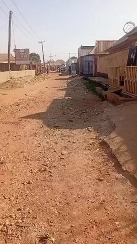 The video shows a dirt road with gravel and stones, typical of a rural or less developed area. The road stretches into the distance, flanked by low buildings and houses on either side.  Power lines are visible overhead, supported by wooden poles.  A sign that says "SPIRIT AT BEST" is visible on the left. A few people can be seen walking in the distance down the road. The weather appears sunny.
