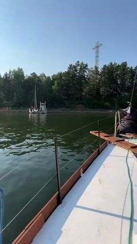 A video of a lake with boats and trees. The sun is shining and the sky is clear. In the foreground, part of a boat is visible, with a rope tied to a railing. In the middle ground, a sailboat is anchored on the water. In the background, a wooded shoreline is visible with some people near the water. A power line tower is also visible on the right side of the screen. The video appears to be taken from another boat moving slowly on the lake.