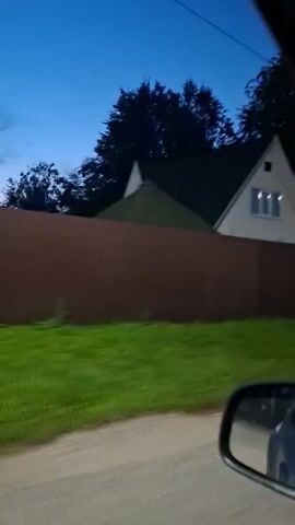 This is a video of a residential house with a pitched roof and multiple windows, partially obscured by a brown wooden fence. The house is light-colored, possibly white or cream, with a dark-colored roof. A section of green grass is visible in front of the fence. The sky appears to be a deep blue, suggesting it is either dusk or dawn. The video seems to be filmed from a moving vehicle, as indicated by the side mirror visible in the bottom right corner and the slight blurriness. There are trees visible behind the house and along the fence line. A power line tower can be seen in the distance on the right side of the frame.