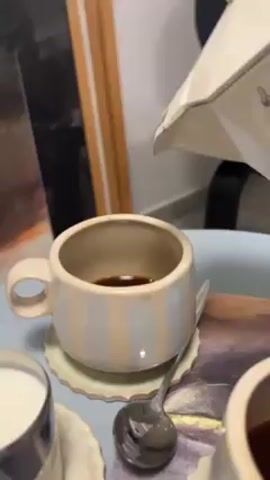 A close-up video shows coffee being poured from a white, Bialetti stovetop espresso maker into a small, vertically striped cup. The cup sits on a light green, fluted saucer. Next to the cup is a silver spoon and a small container filled with milk. The background includes framed artwork and a table.
