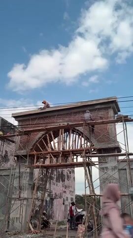 This is a vertical, eye-level shot of an arched gateway under construction in Klatakan, Indonesia. The sky above is a bright blue with scattered white clouds, suggesting it is a sunny day, likely in the morning or afternoon.

The main subject is the unfinished brick archway, supported by extensive scaffolding made of metal poles and wooden planks. The arch itself is ornate, with decorative brickwork and what appears to be wooden framing for the curved roof. Workers are visible on and around the structure, engaged in the construction process. Two individuals are perched on top of the arch, one seemingly working on the horizontal beam, while another is on the right side of the structure, also appearing to be working. Closer to the ground, two other people are visible near the base of the arch, one standing and looking up, and another sitting. In the middle ground, a group of three people are gathered, with one person in a red shirt holding a tool.

To the left of the archway, a section of a weathered building with peeling pink paint is visible, adding a sense of age and history to the scene. The ground appears to be dirt and construction debris. Power lines are strung across the top of the frame, indicating the presence of infrastructure in the area. The overall atmosphere is one of ongoing labor and development.