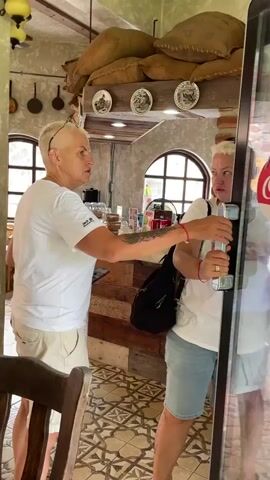 Two people are standing in front of a glass door, which has an "Open" sign on it. One person, who appears to be a woman with short blonde hair, is wearing a white t-shirt and tan shorts. She is reaching out to the other person's hand, which is on the door handle. The second person, also appearing to be a woman, is wearing a white t-shirt and denim shorts. She is holding a green bottle in her other hand. The environment appears to be a cafe or shop, with sacks of coffee beans visible in the background. The floor has a patterned tile design.