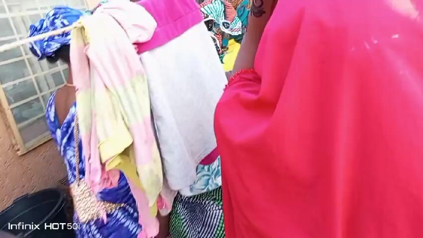 A group of women and children are gathered outside, likely at a celebration or event. The women are wearing colorful traditional clothing, including headscarves, and some are seated on the ground. There are some clothes hanging on a rope, obscuring the view in the initial frames. People appear to be watching something, possibly a performance or a ritual. Several people are recording with their phones. The environment seems to be a residential area.