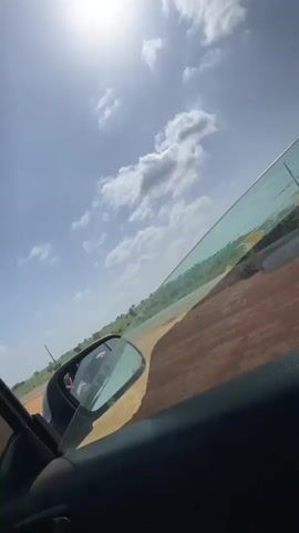 The view from inside a moving vehicle presents a slightly tilted frame, capturing a landscape under a bright, sunny sky with scattered clouds.  The vehicle's side window is partially visible, tinted with a subtle green hue.  Outside the window, a predominantly brown and tan landscape stretches, punctuated by sparse greenery.  The side mirror of the car is also visible, reflecting the interior of the vehicle and a hint of the driver.  The sun shines brightly in the sky, casting a light glare.