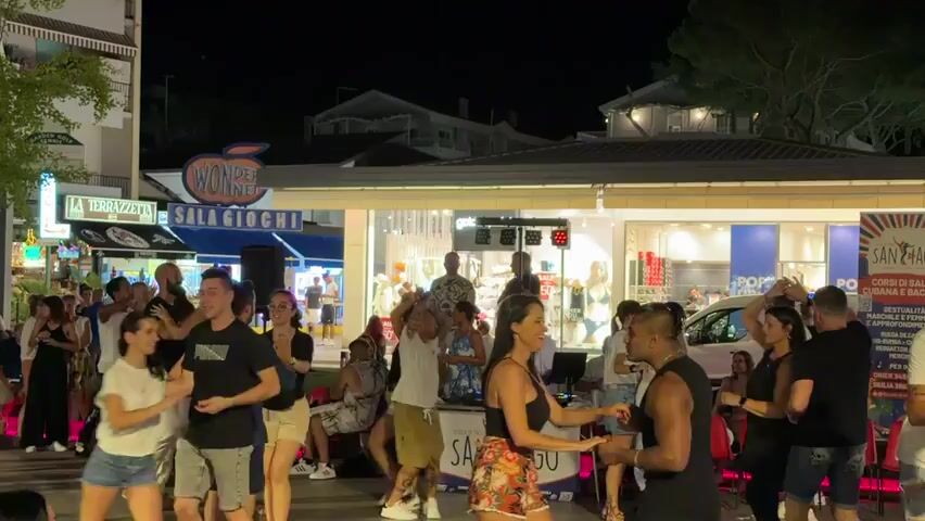 The video shows a crowded outdoor scene at night, possibly a street or plaza, where a group of people are dancing. In the foreground, a man and a woman are dancing closely together, with the man wearing a black tank top and the woman in a black top and a floral skirt. To their left, a group of people are also dancing, some in pairs. To the right, more people are gathered, watching the dancers or standing around. Behind the dancers, there are shops and buildings illuminated by artificial lights. A sign with a red apple logo and the words "WONDE SALA GIOCHI" is visible above one of the shops. There is a DJ booth with lights and equipment in the middle of the dancing area. The overall atmosphere appears to be lively and festive.