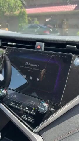 A close-up shot of a car's infotainment system displays a song playing via Bluetooth 5.1. The screen shows the artist as Ernest Oganesyan and the song title as Sevani Apin, with album art of a woman in a traditional dress. The car's climate control displays a temperature of 22.0 degrees Celsius for both driver and passenger sides. The background is slightly blurred, showing the exterior of a building with a red roof and a parked car. A person in a white t-shirt walks into the frame from the left and walks away from the camera. The timestamp indicates the video begins at 00:00 and ends at 00:05.