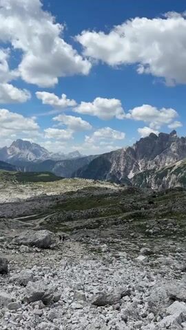 The media file presents a panoramic view of a rugged mountainous landscape under a bright, partly cloudy sky. The foreground is dominated by a scree slope, a chaotic jumble of large, grey rocks interspersed with patches of green grass. In the mid-ground, the terrain becomes more vegetated, with rolling hills and further rocky outcrops. In the distance, majestic, jagged mountain peaks rise, their slopes a mixture of rock and verdant green, some areas appearing to be snow-capped. The sky is a vibrant blue, dotted with fluffy white clouds that cast shadows on the mountainside, suggesting it is midday.

Two small figures, appearing to be hikers, are visible on a path winding through the mid-ground, suggesting human presence and activity in this remote wilderness. The overall mood is one of awe-inspiring natural beauty and a sense of adventure.

A notable detail is the word "MESSI" written in large white letters on a grassy slope in the mid-ground, made of possibly stones or painted markings. This text stands out against the natural surroundings, hinting at a playful or symbolic gesture within the vast landscape. The location is identified as Toblach - Dobbiaco, Italy, a region known for its stunning Dolomite scenery.