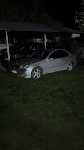 The video presents a dimly lit outdoor scene at night. Three cars are parked under a shelter with a corrugated metal roof supported by several metal poles. The cars appear to be parked on a concrete surface, with grass visible in the foreground. The closest car to the camera is a silver Mercedes-Benz, followed by a dark-colored BMW with its headlights on. A third, darker vehicle is parked further back. The cars have license plates, but the details are difficult to discern. The overall atmosphere is dark, with some red lighting reflecting off the roof of the shelter in the background.