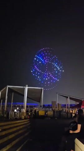 A drone light show is taking place at night. The drones are forming various shapes in the sky, illuminated with blue and purple lights. Below the drone display, there is a crowd of people gathered, with some standing behind metal barricades. Structures with white canopies are visible in the foreground, and there are lights and possibly stage equipment set up. The overall environment suggests an outdoor event or performance.