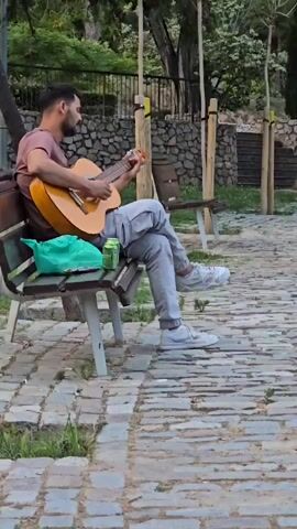 A man sits on a park bench playing an acoustic guitar. He is wearing a pinkish-brown t-shirt, grey cargo pants, and white sneakers. A green bag and a can are on the bench next to him. The bench is on a paved walkway in a park. In the background, there is a stone wall and trees.
