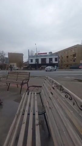 The video shows an outdoor urban scene on a cloudy day.

A young man with dark hair, wearing a brown jacket, black pants, and brown shoes, walks across the sidewalk, looking at something in his hands. He passes by a decorative lamppost and a red, glass-walled ATM enclosure. Another person can be seen inside the ATM enclosure. 

The background features benches lined up along the sidewalk, a street with moving traffic, and buildings. One building has a sign in a language other than English that reads "SAMARKANDOR." The ground appears wet, suggesting recent rain. The overall atmosphere is somewhat bleak due to the overcast sky.