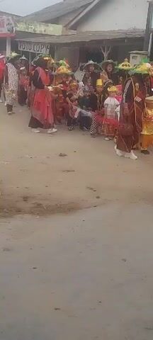 This video shows a parade of people, including children, walking down a dirt road. The participants are wearing colorful traditional clothing and hats. The parade appears to be a cultural event or celebration. The background shows buildings and signs, suggesting an urban or semi-urban setting. Some individuals are holding umbrellas or parasols, which are decorated with colorful flowers. The overall atmosphere seems festive and joyous. The video is shot from a slightly elevated angle, looking down at the parade.