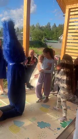 A group of children are gathered outdoors on a wooden porch. One child, dressed in a full-body blue furry costume with a large headpiece, is kneeling on a patterned mat. The costumed figure is holding a cake with candles. Several children are watching, some clapping. One girl in a white dress is standing close to the costumed figure. Another girl in pajamas is facing the costumed figure. The environment includes wooden porch railings and a blue sky with clouds.